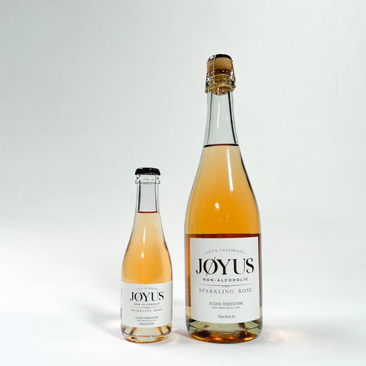 Two Joyus sparkling wine bottles, one larger bottle and one smaller bottle, filled with pale pinkish-amber wine, standing on a white studio background.