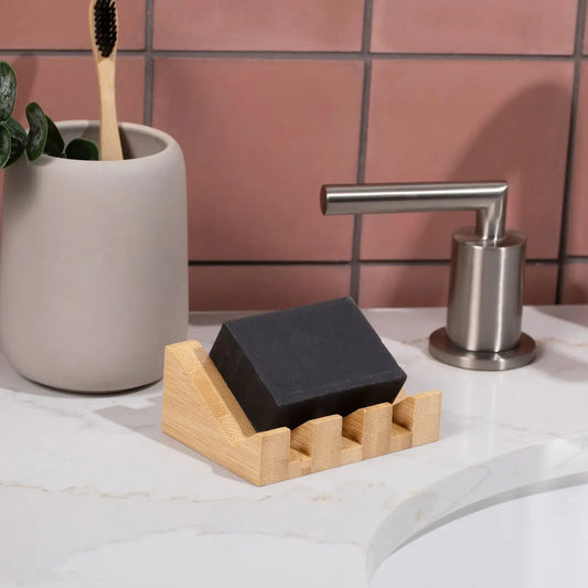 Black soap bar on a bamboo soap dish with a plant and soap dispenser in the background.