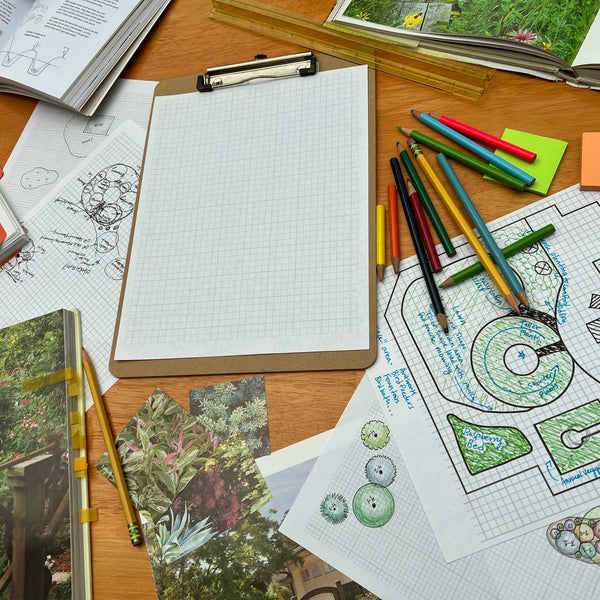 A table covered in graph paper landscape design sketches, colorful pencils, books, and pictures of beautiful garden plants... Representing the landscape design consultation raffle item