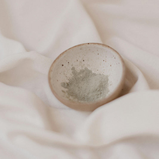 A small bowl of French Green Clay powder, placed on a light fabric surface.